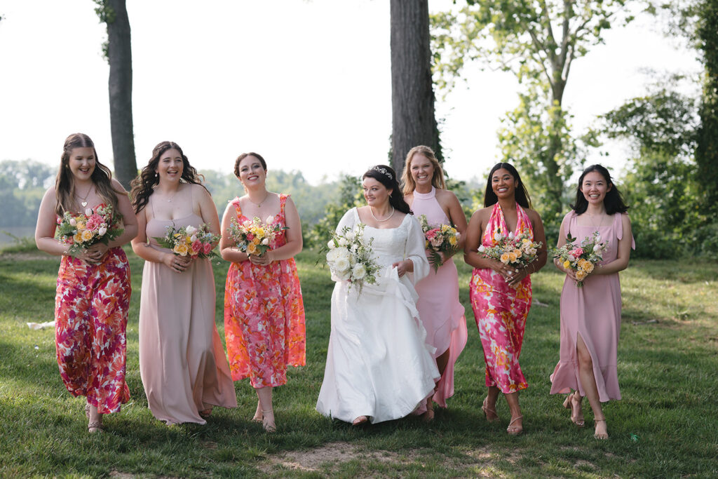 A bride and her bridesmaids walk together outdoors, smiling and holding bouquets of flowers. The bride wears a white dress, while the bridesmaids are dressed in shades of pink and floral prints, creating a vibrant and joyful scene against a green, natural backdrop.