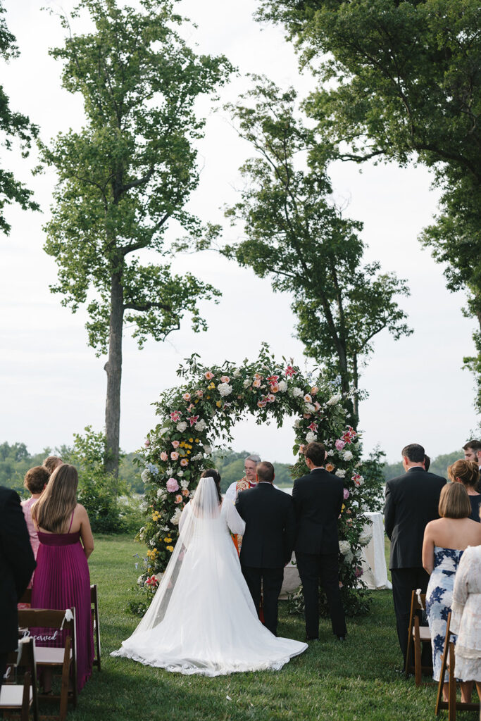 A wedding ceremony taking place outdoors, viewed from behind the bride and groom who stand before a floral arch adorned with colorful flowers. The bride wears a long white gown with a veil, and the groom is in a black suit. Guests are seated on either side, watching the couple exchange vows against a backdrop of tall trees and open sky.