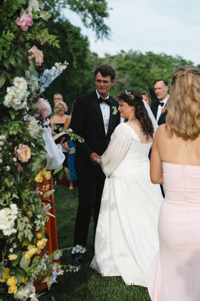 A bride and groom stand together during their wedding ceremony, smiling as they hold hands. The bride wears a white dress with a veil and tiara, while the groom is in a black suit with a bow tie. They are surrounded by guests and a lush floral arrangement, creating a picturesque outdoor setting.