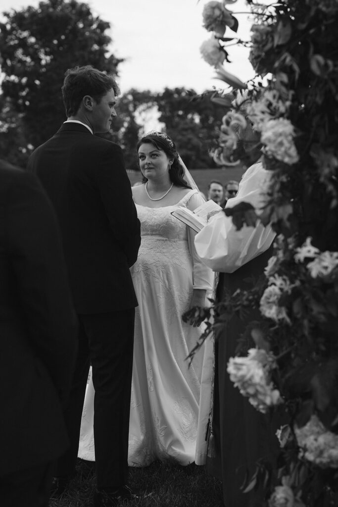 A black-and-white photo of a bride and groom during their wedding ceremony, facing each other as they exchange vows. The bride, wearing a white dress and a tiara, looks at the groom with a serene expression.