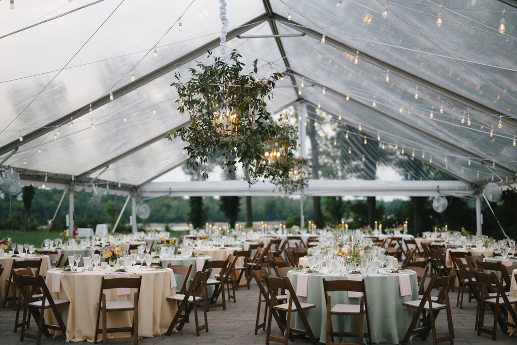 A beautifully decorated wedding reception under a clear tent, featuring round tables draped in pastel-colored linens and set with elegant glassware and floral centerpieces. Overhead, string lights and greenery-adorned chandeliers add a warm and romantic ambiance to the spacious outdoor setting.