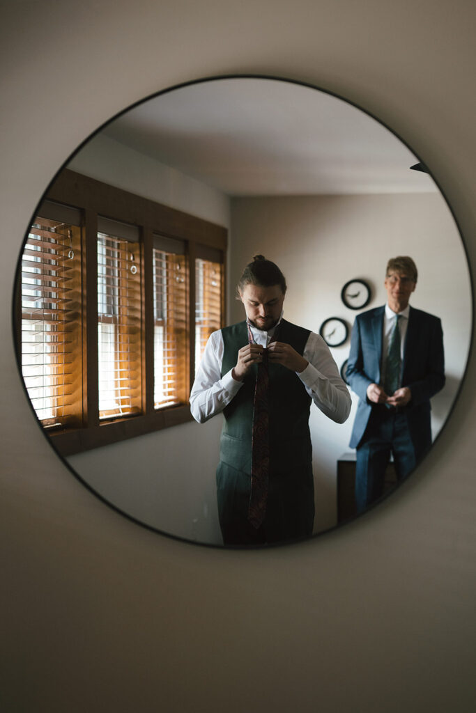 A groom, reflected in a circular mirror, is adjusting his tie while his groomsman stands behind him. They are in a well-lit room with wooden blinds and wall clocks.