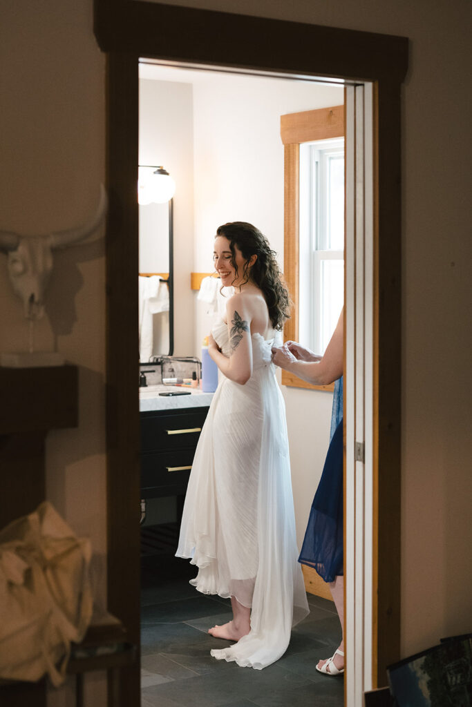 A bride, smiling and standing barefoot, has her wedding dress adjusted by another woman in a blue dress. The scene is set in a warmly lit room with wooden accents and a vanity in the background.