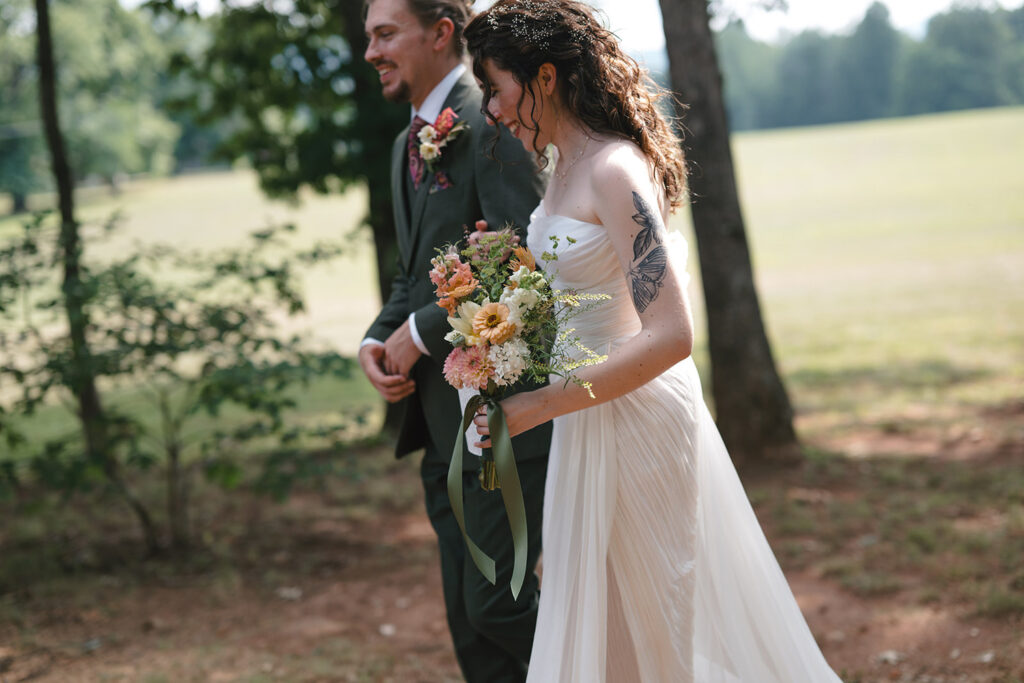 A bride and groom walk arm in arm outdoors, smiling and surrounded by greenery. The bride carries a bouquet of colorful flowers and wears a white dress, while the groom wears a dark suit adorned with a boutonniere.