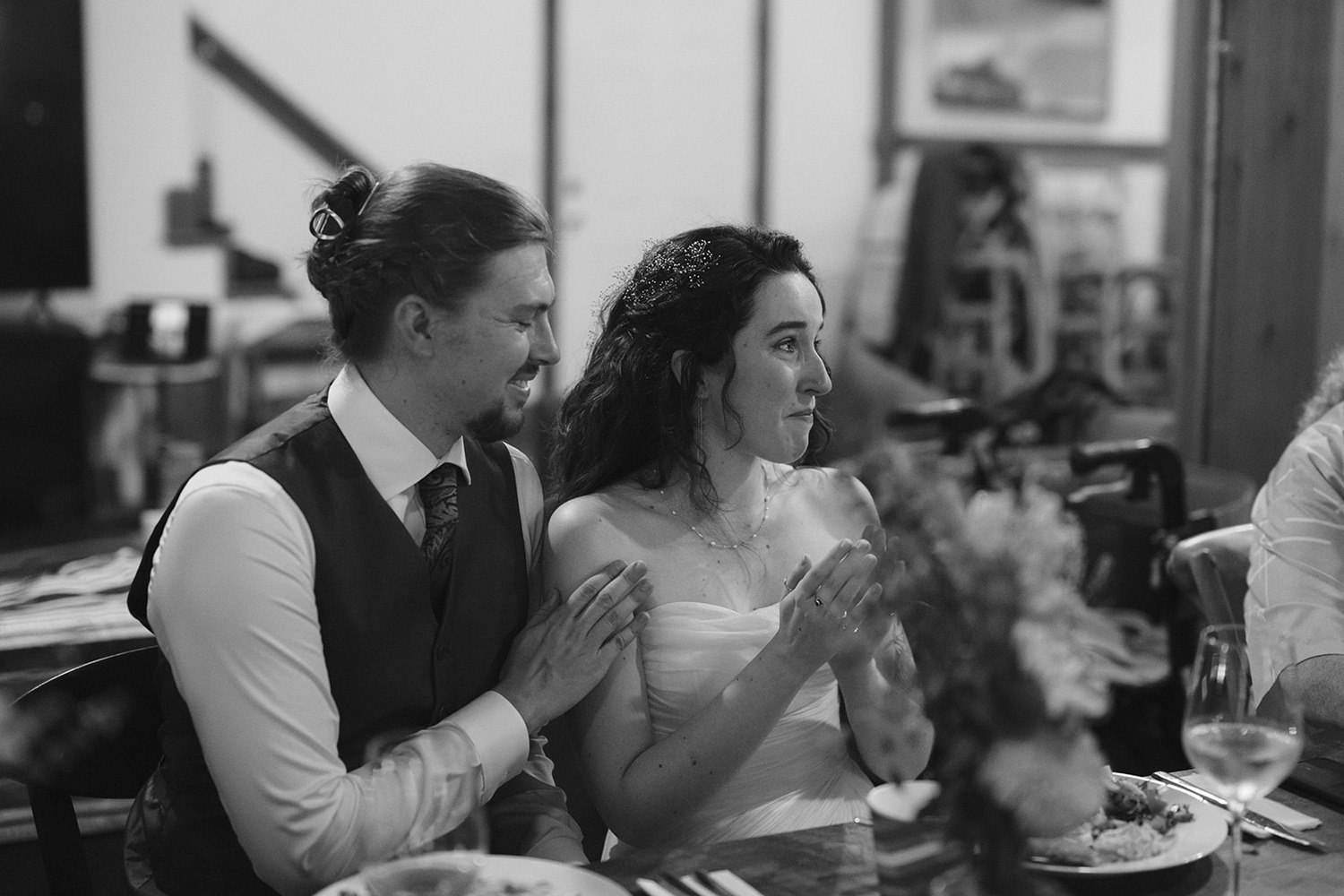 A bride and groom sit together at a reception table, smiling and applauding. The bride, in a strapless dress, looks touched and emotional, while the groom, wearing a vest and tie, gently places his hand on her shoulder. The setting is warm and intimate, with food and wine on the table.