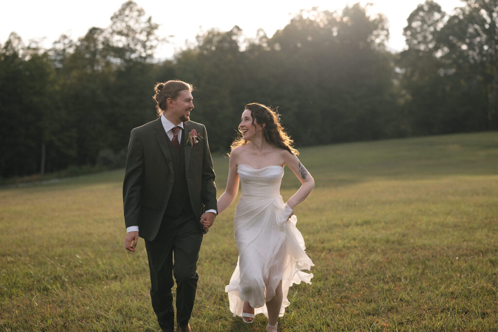 A bride and groom hold hands and walk through a sunlit field, smiling at each other. The bride wears a flowing white dress, and the groom is dressed in a dark suit. The backdrop features trees and a serene, natural setting.