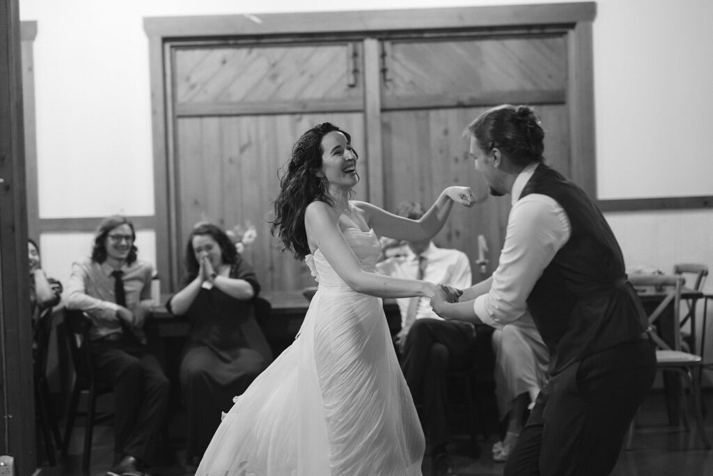 A bride and groom dance joyfully in a rustic indoor setting, while seated guests in the background watch and cheer with smiles and applause. The bride's dress flows gracefully as they move.