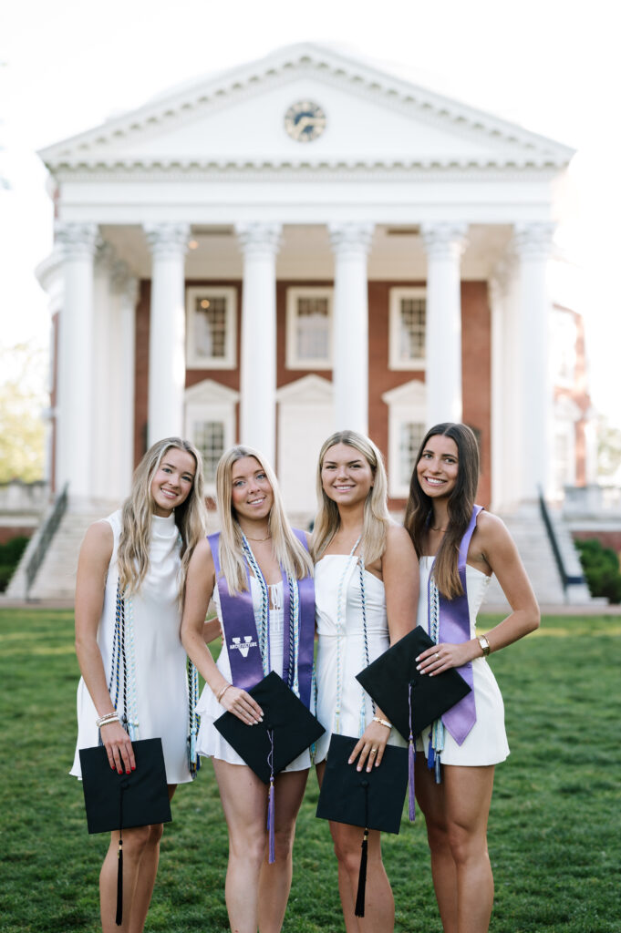 4 women in white smile at the camera while holding graduation caps on the lawn of university of virginia's campus