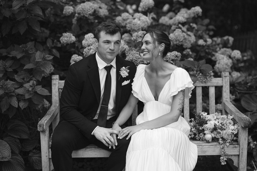 Black and white photo of a newlywed couple sitting on a bench holding hands 