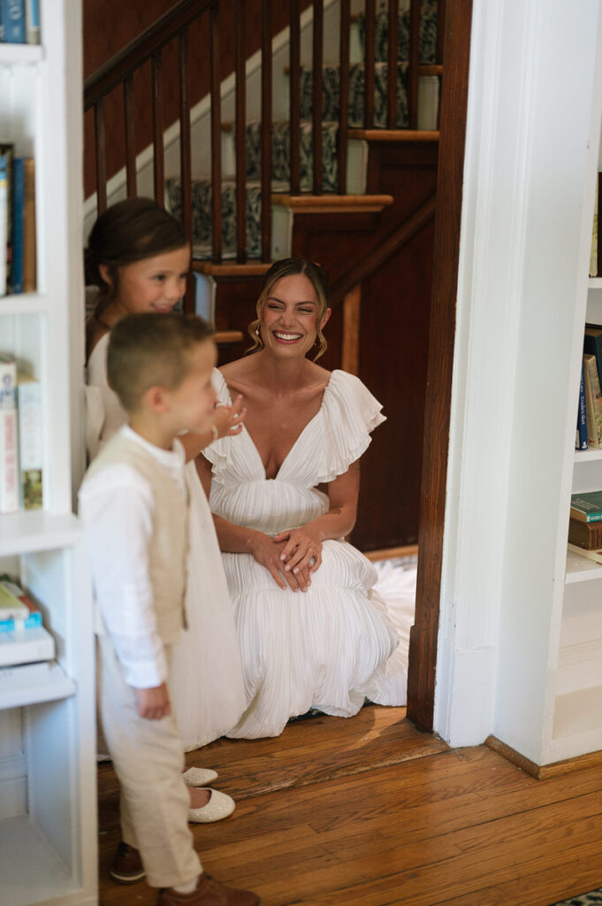 A person in a wedding dress smiling and crouching down with two small kids in front of them