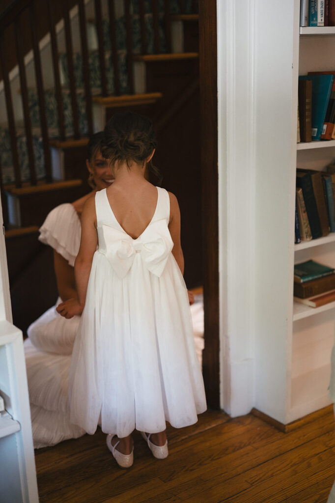 A young girl in a white dress with a large bow on the back standing facing a person in a wedding dress 