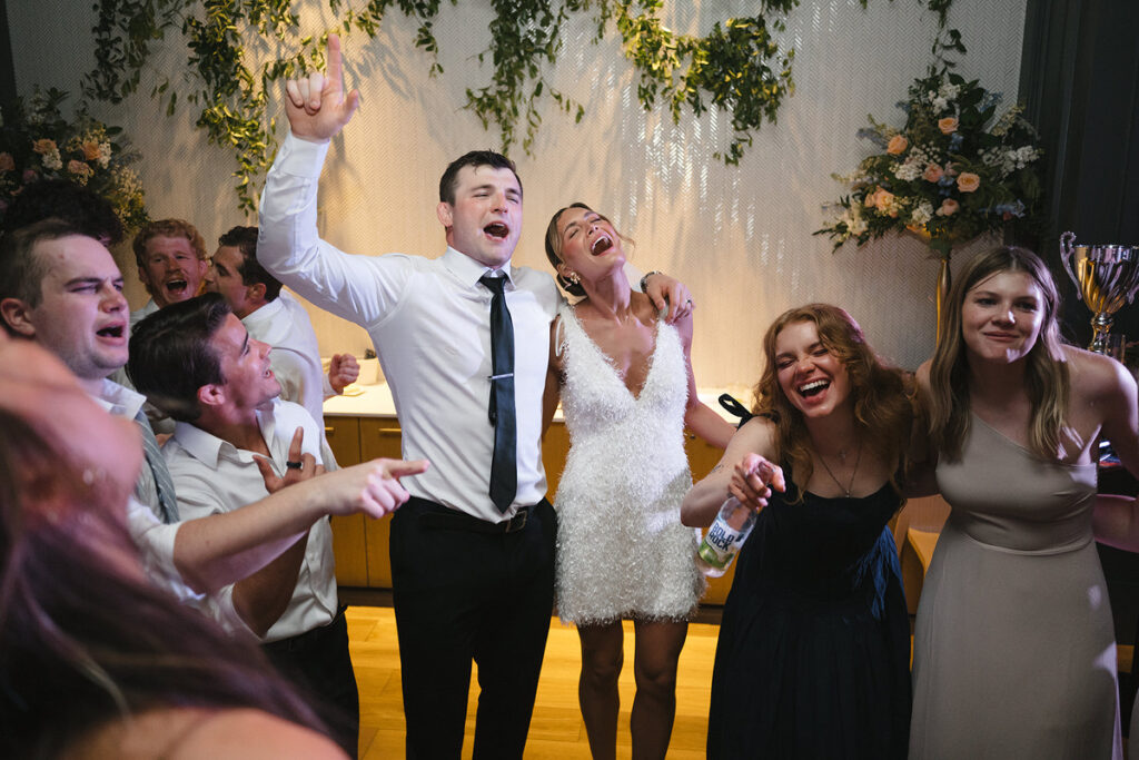 A newlywed couple and their wedding guests dancing and singing. 