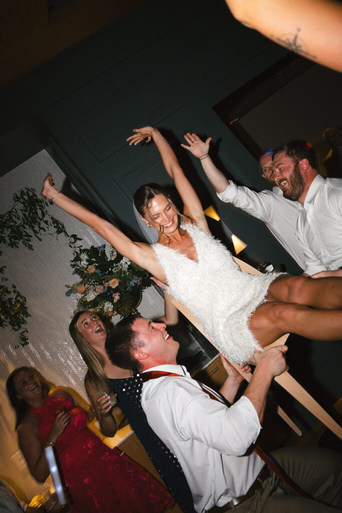 A newlywed being lifted up in a chair while wedding guests dance around them 