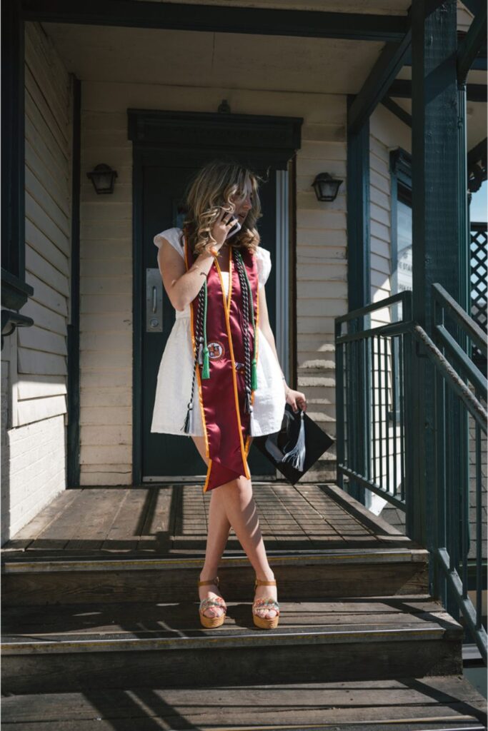 A woman in graduation stoles standing on a porch, talking on the phone while holding her graduation cap, wearing wedge sandals and a white dress.