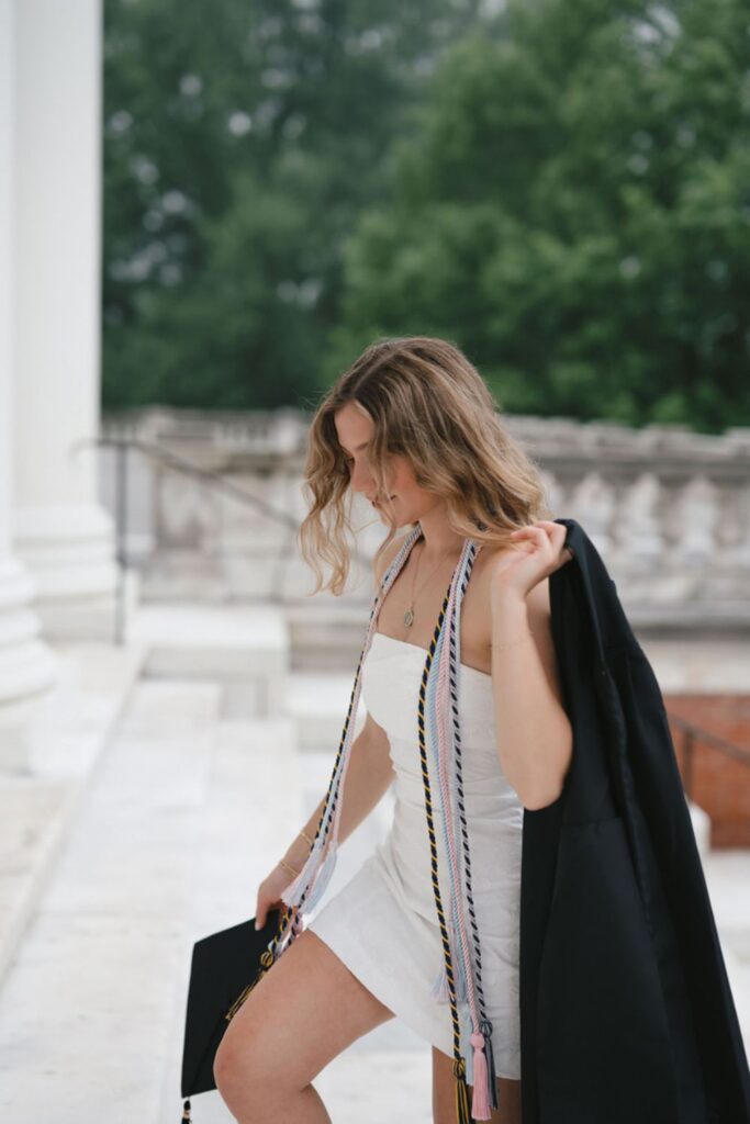  A woman wearing a white dress and several honor cords, walking down marble steps, holding her graduation cap in one hand and a black jacket in the other.