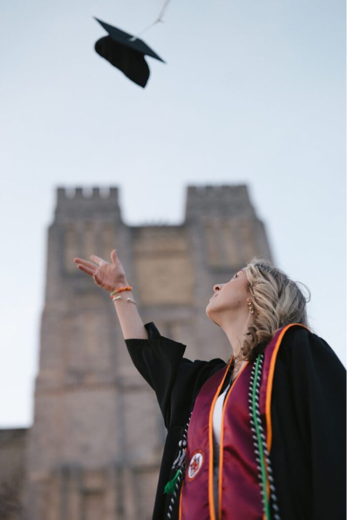 A woman wearing graduation stoles and a black gown throws her graduation cap into the air, with a large stone building in the background, symbolizing the celebration of her achievement.