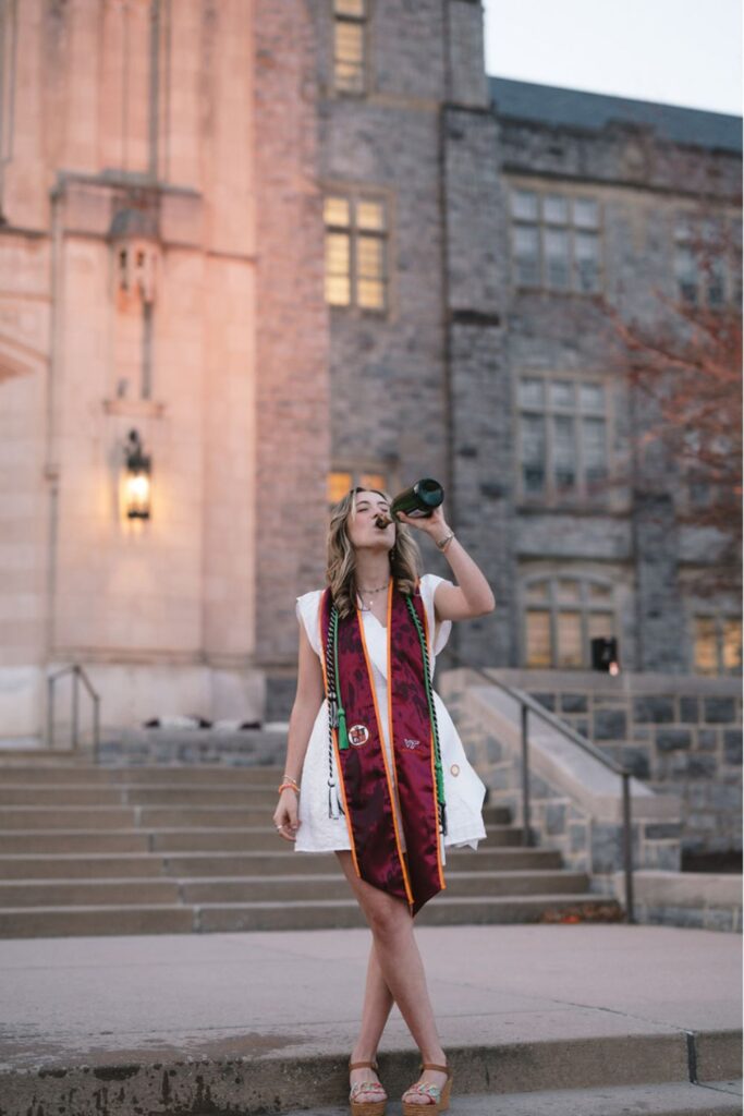 A woman in graduation stoles and a white dress, standing on steps in front of a stone building, drinking from a bottle of champagne.