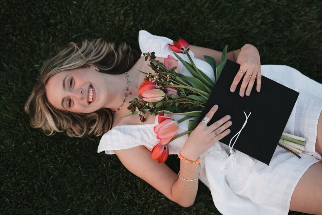 A woman in a white dress lying on green grass, holding a bouquet of tulips and her graduation cap, smiling happily.