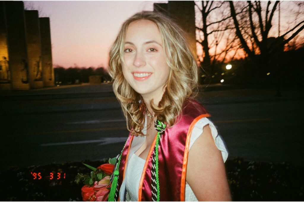 A woman smiling at the camera at sunset, wearing graduation stoles and holding a bouquet of pink flowers, with a soft orange sky in the background.