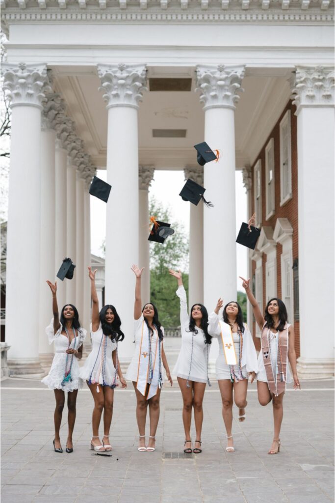 Six women in white dresses and graduation stoles, tossing their caps into the air beneath a grand structure with tall columns, smiling as they celebrate.