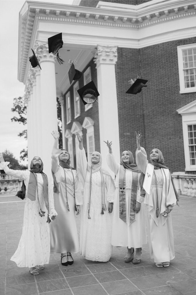 Five women in graduation attire, throwing their caps in the air in celebration in front of a brick building with large white columns.