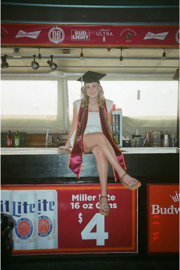 A woman in graduation attire, sitting on a bar counter with a smile, surrounded by beer advertisements in a casual environment.