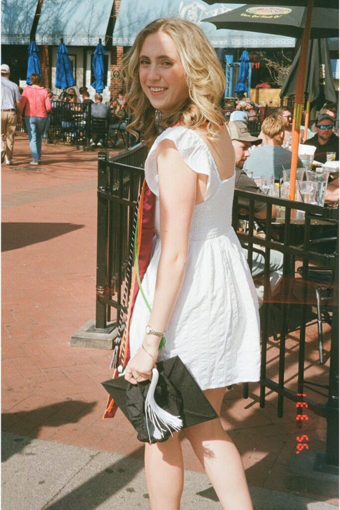 A woman in a white dress walking outside, holding her graduation cap and smiling, while people sit at a café in the background.