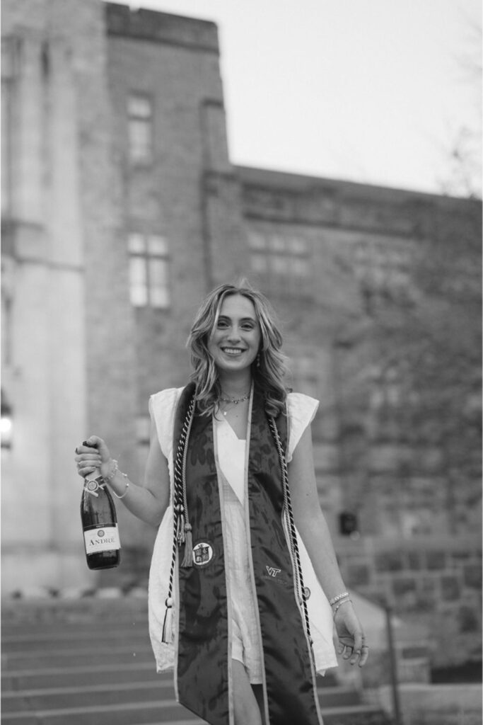 A smiling woman holding a bottle of André champagne and wearing graduation stoles, posing in front of a university building made of stone.