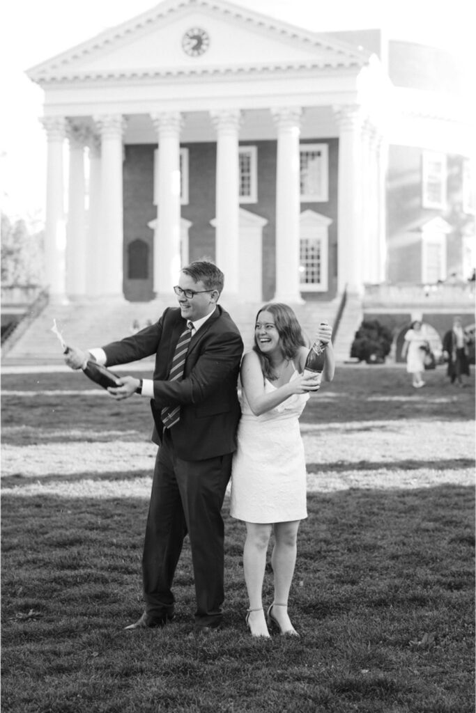 A man and a woman, both dressed in formal attire, enthusiastically popping champagne bottles on a lawn, with a large building featuring columns in the background.