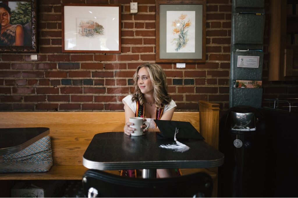 A woman sitting alone in a café, holding a coffee cup and looking out thoughtfully, with her graduation cap resting on the table in front of her.