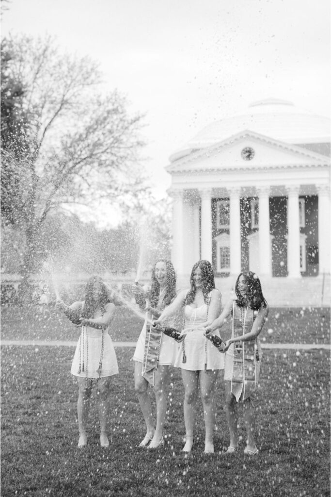 Four women dressed in white, joyfully popping champagne bottles on a lawn in front of a university building with columns, with champagne spraying in the air.