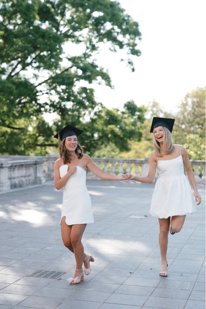 Two women in white dresses and graduation caps, laughing and holding hands while walking down a stone terrace with greenery in the background.