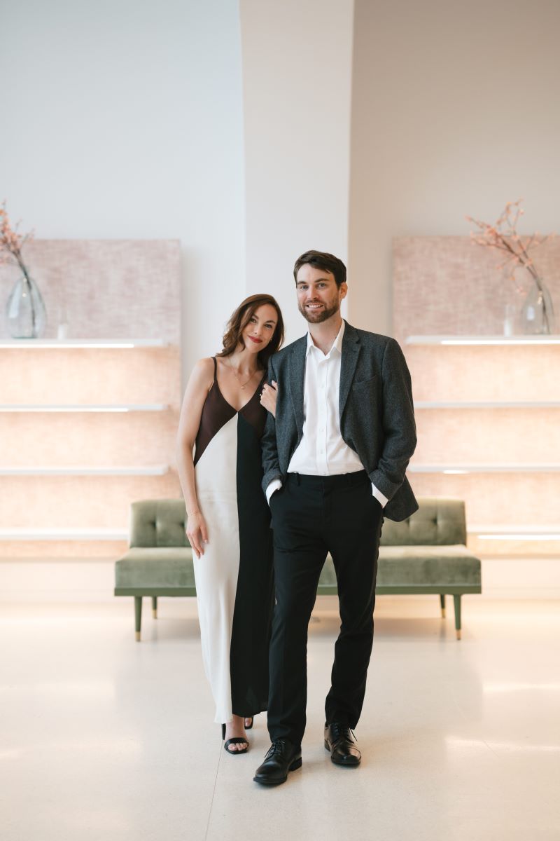 A man in a suit with a white button up and a woman in a black and white dress smiling the woman is grabbing the arm of the man and the man has his hands in his pockets behind them is a green sofa 