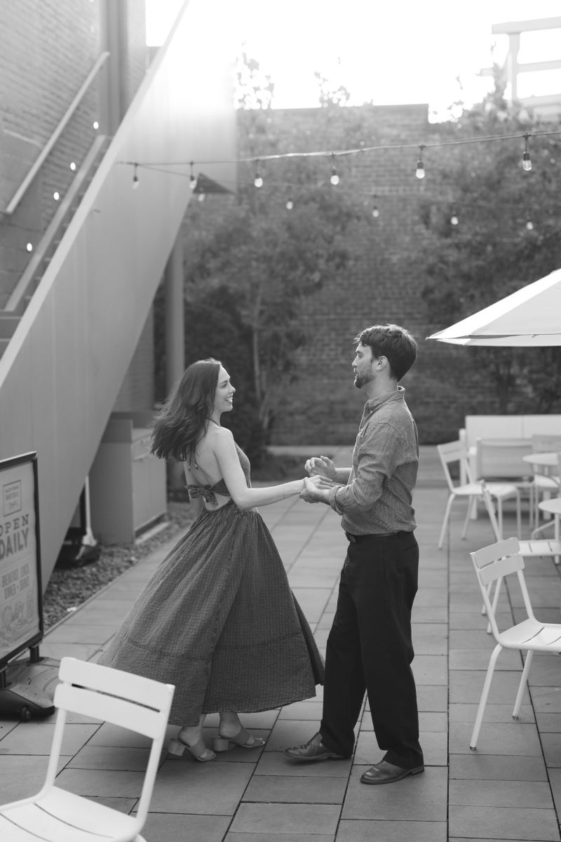 A man and woman on a patio with string lights, tables, and chairs the man is dancing with the woman who is wearing a dress and heels and the man is wearing a button up and slacks 