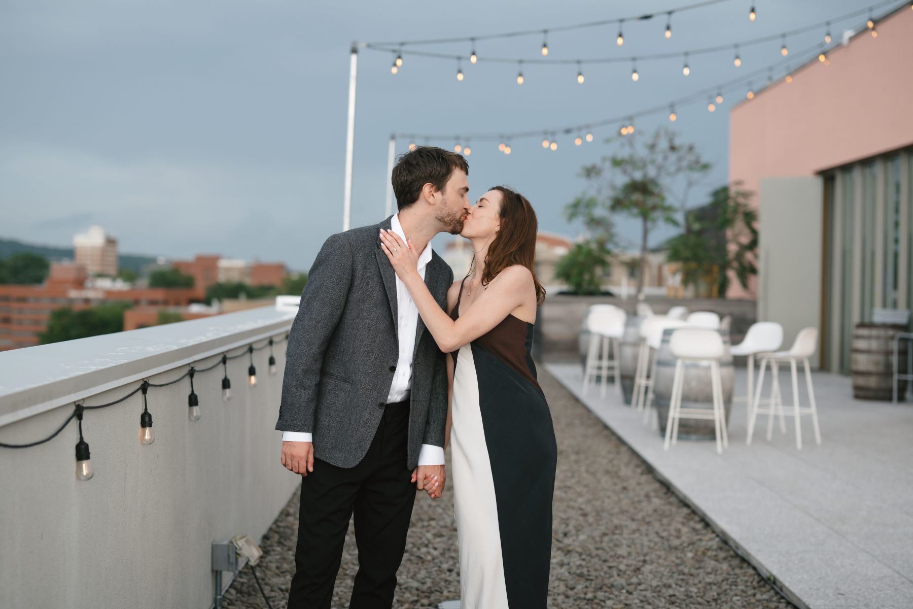 A man and woman kissing on a rooftop with string lights and tables and charis the woman is wearing a black and white dress and touching the man's shoulder and the man is wearing a gray blazer and white button up and black pants the couple is holding hands 