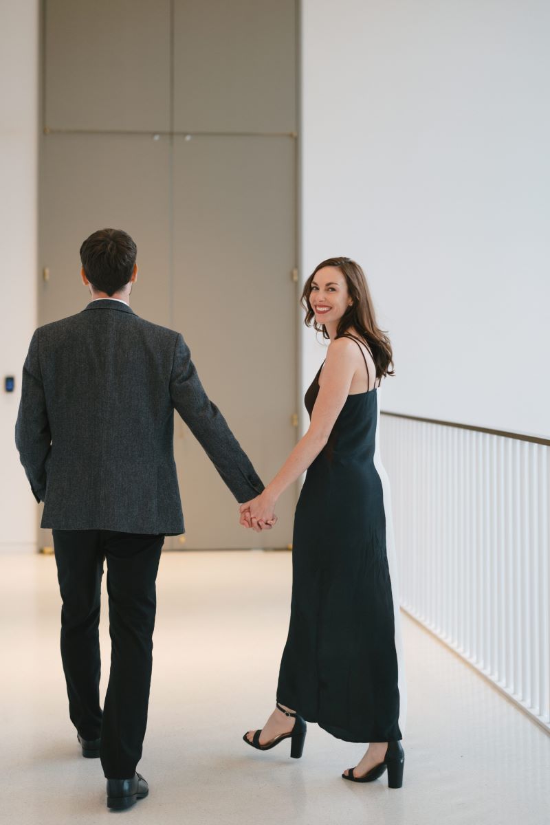 A woman in a black and white dress and heels in holding the hand of a man in a suit they are walking down a hall and the woman has turned back and looked over her shoulder and is smiling 