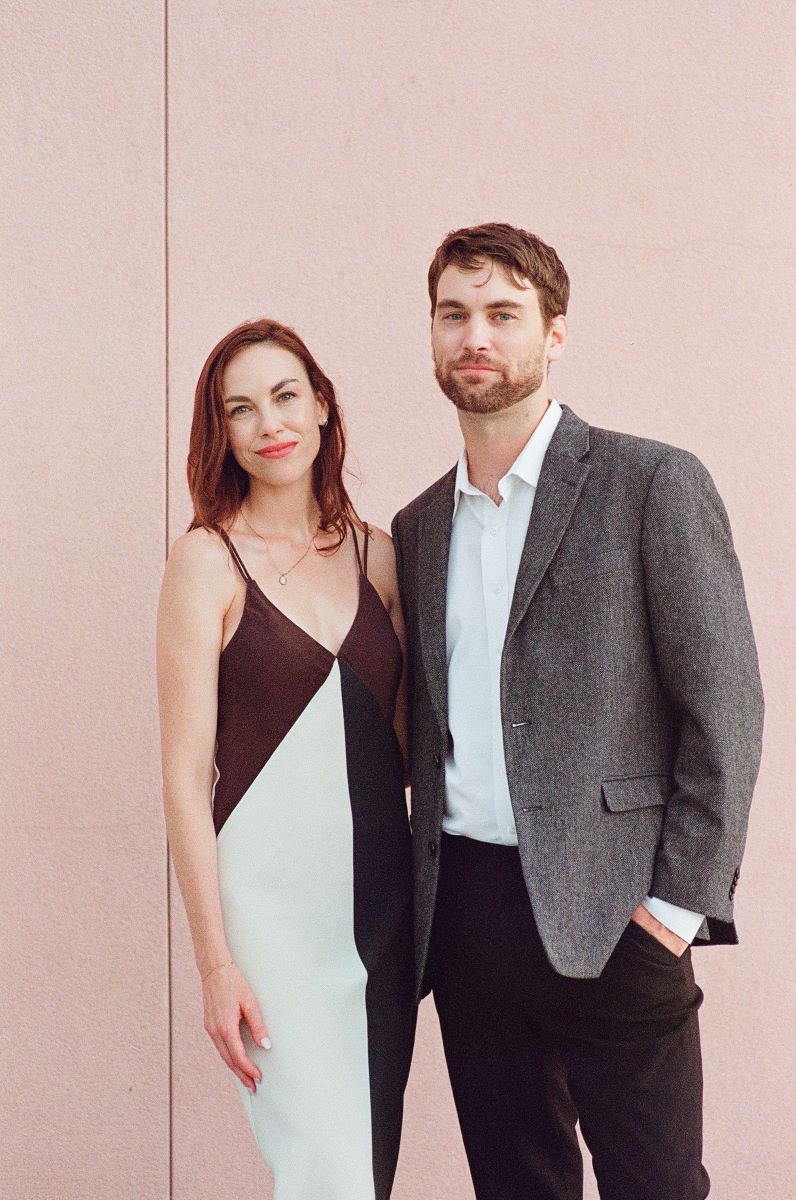 A man in a gray suit wiht a white button up and a woman in a black and white dress and both softly smiling behind them is a plain pink wall 