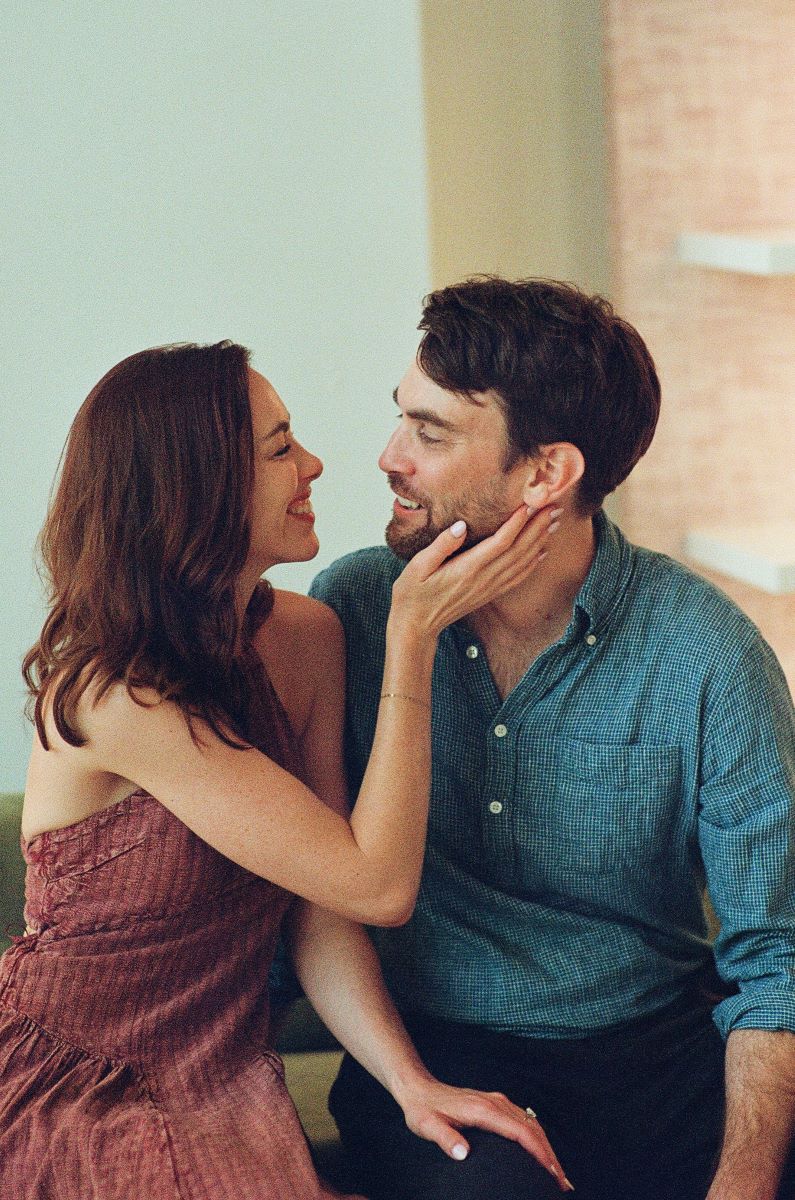 A man in a blue button up is smiling at a woman in front of him the woman is wearing a red dress and is also smiling andn touching the man's face they are both sitting down 