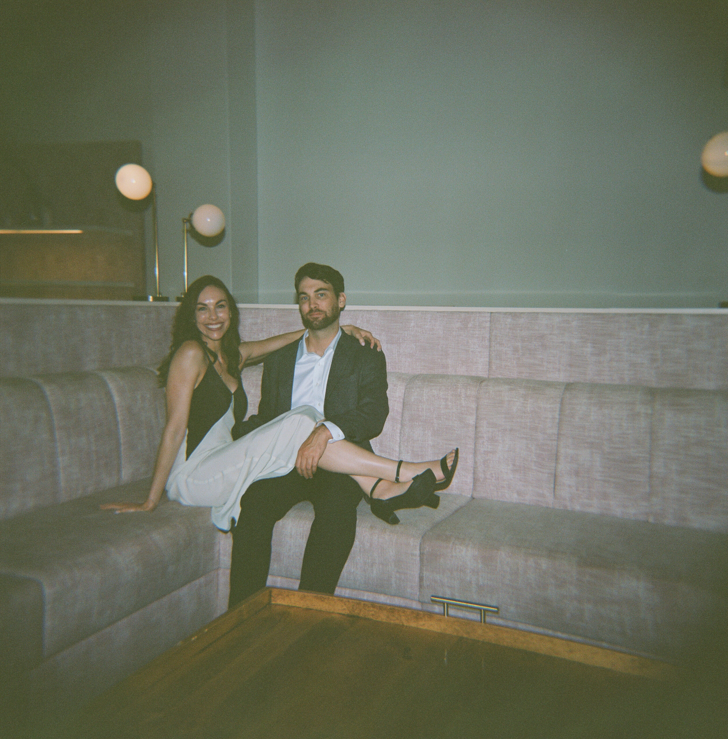 A woman and man sitting on an l-shaped couch the woman has her legs over the man's legs and her arm around his shoulder the woman is in a black and white dress and the man is in a suit with a button up 