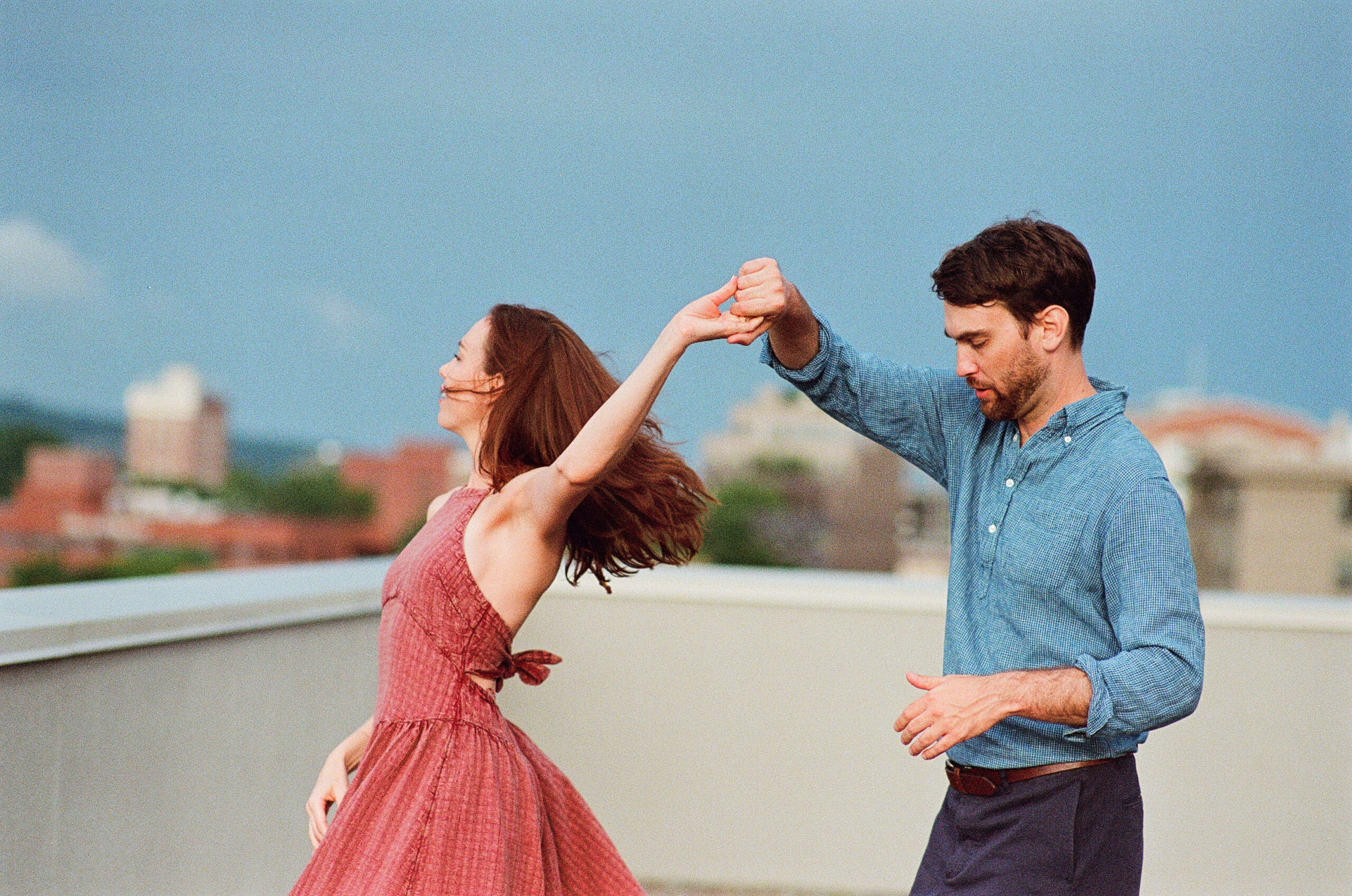 A man in a blue button up and navy pants twirling a woman in a red dress they are both on a rooftop 