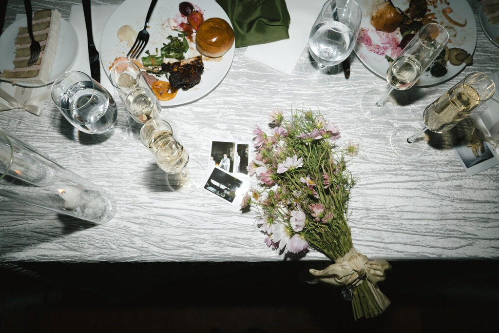 A reception table with half eaten food, champagne drinks, a floral bouquet, and polaroids 