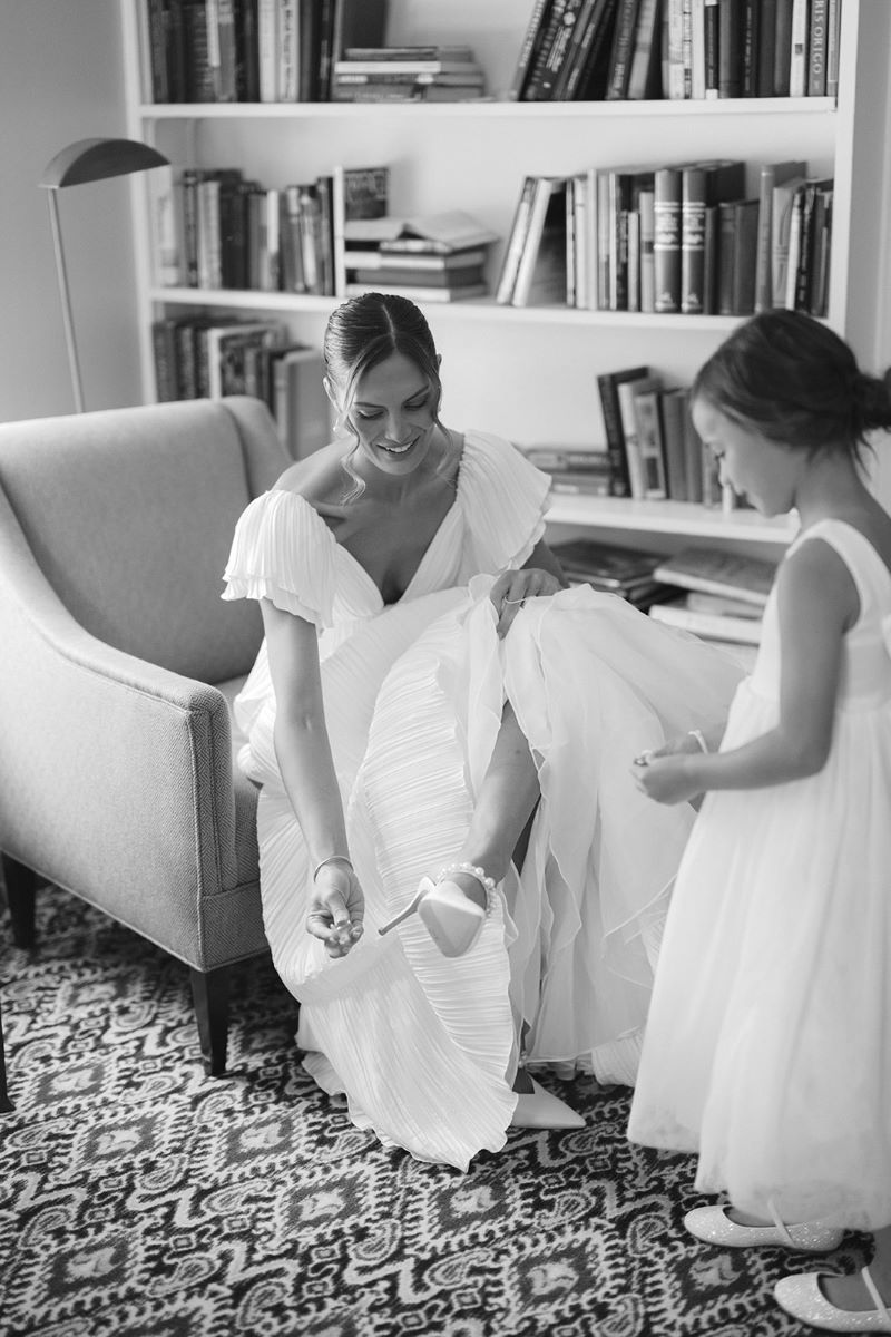 woman fixing her shoe in a wedding dress sitting on a chair with a young girl in a dress standing in front of her 