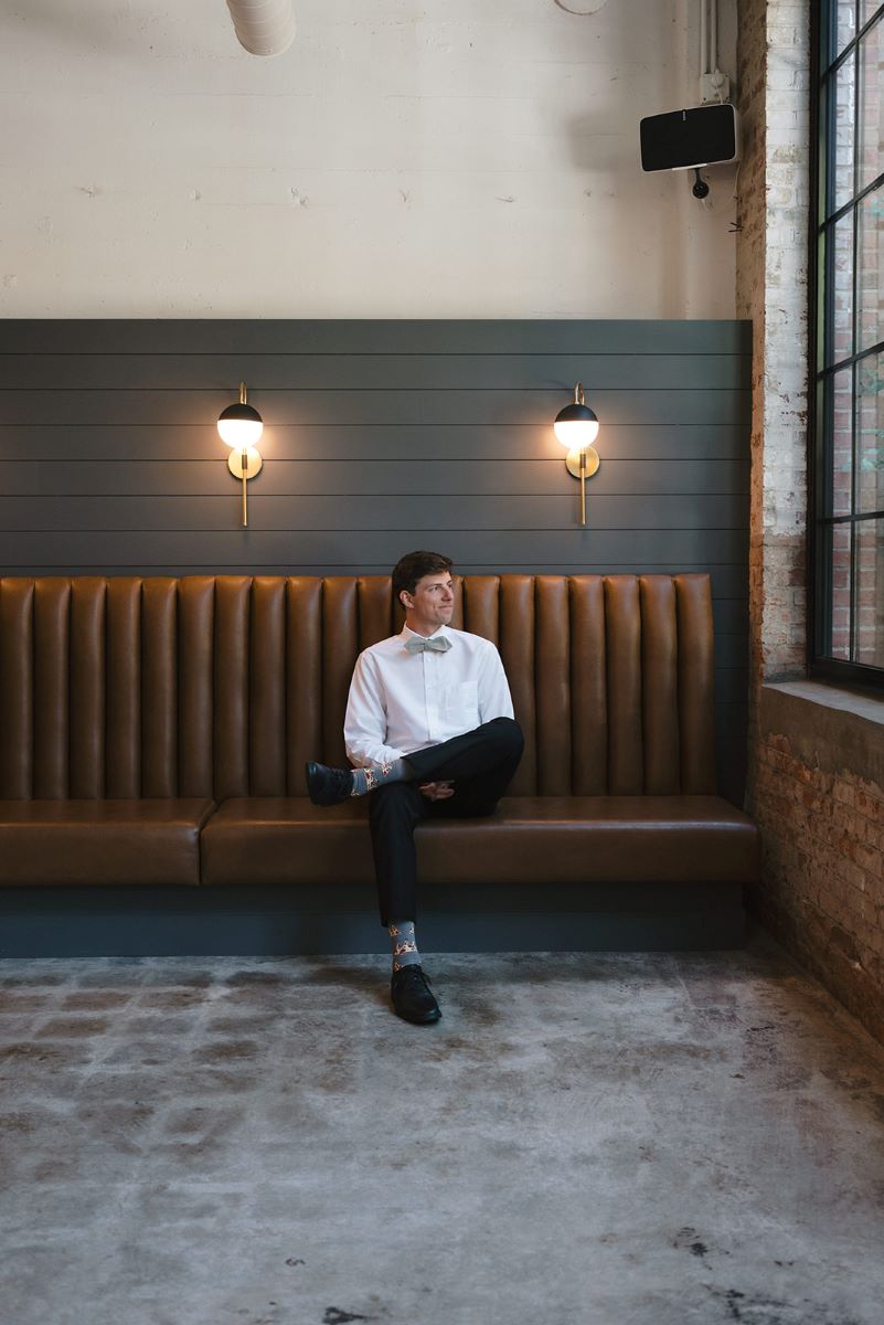 man in a white button-up, black pants sitting on a brown couch looking out a window 