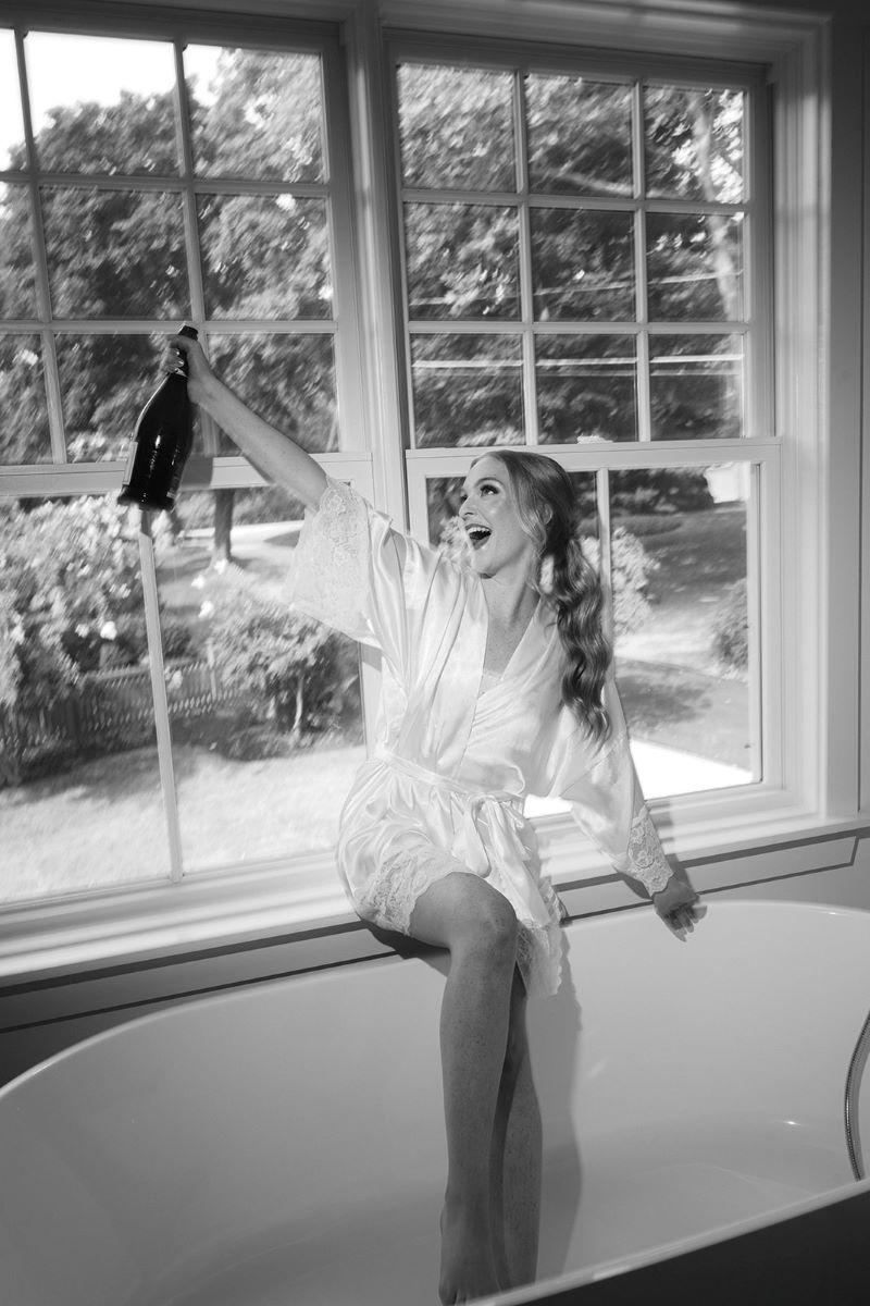 black and white photo of a woman sitting on a window ledge holding a bottle of champange 