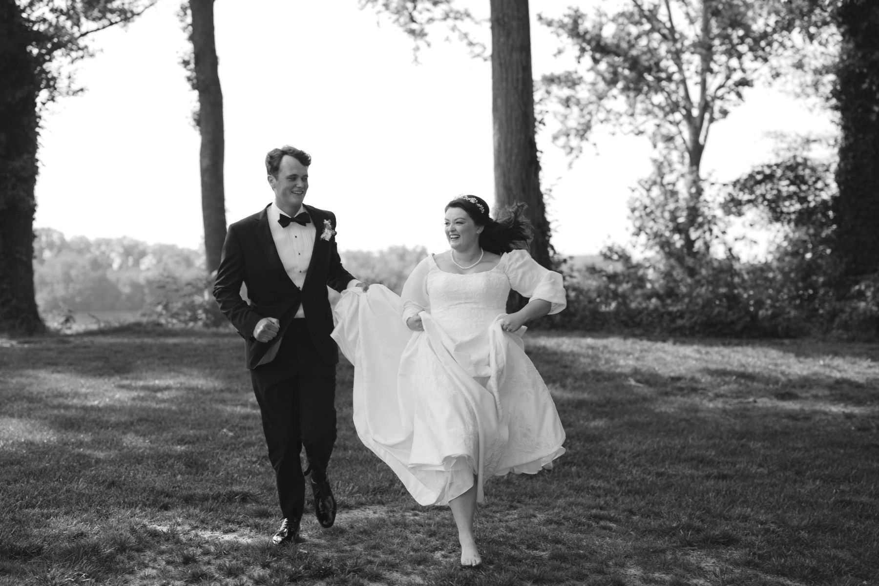 A woman in a white wedding dress wearing a pearl necklace is running through the grass with her partner who is wearing a black tuxedo, a bowtie, and a white button up and he is running throught he grass with her and holding part of her wedding dress 