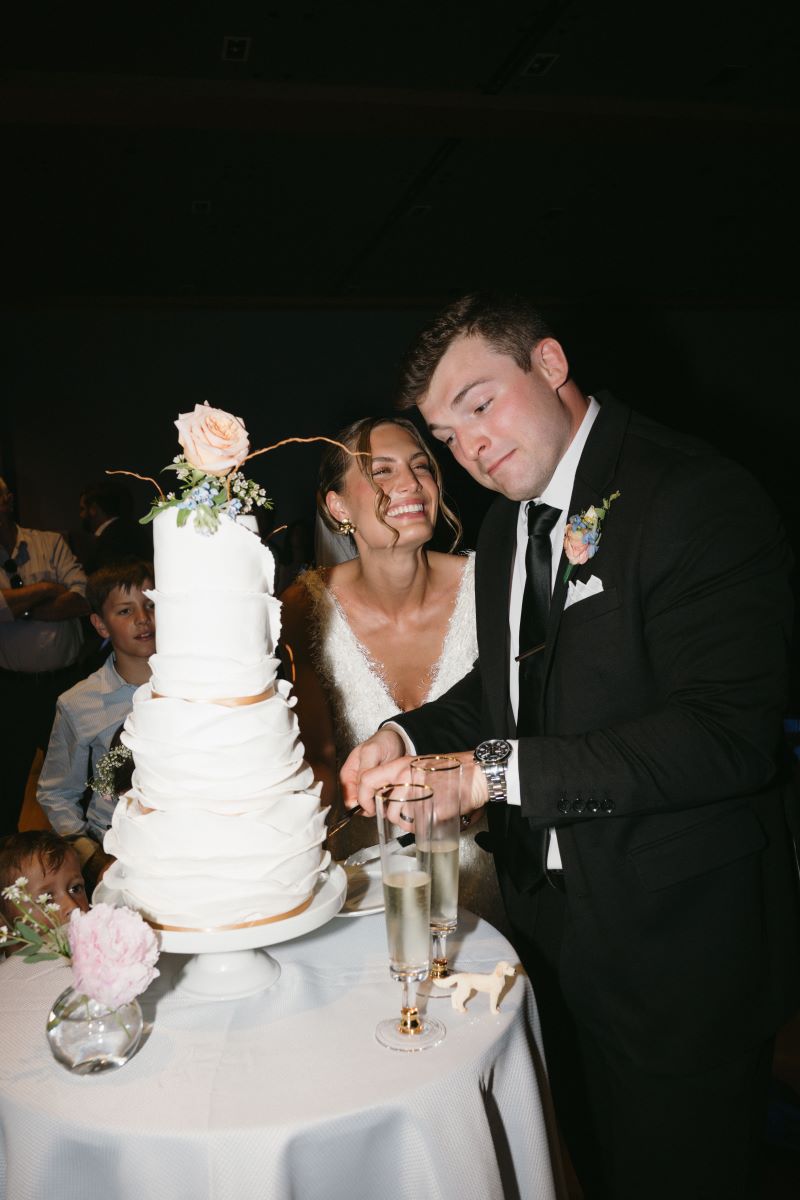 A woman in a white dress is laughing as her partner who is wearing a black tuxedo cuts their wedding cake