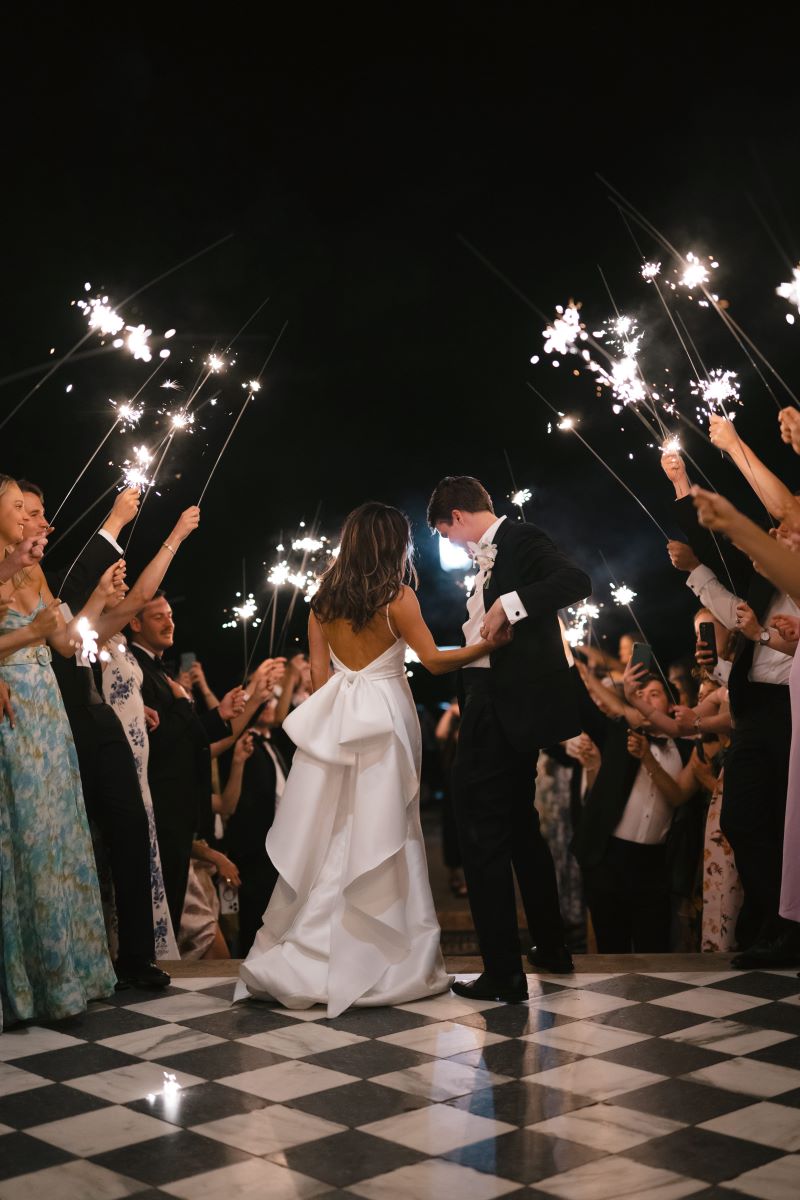 A man in a black tuxedo is dancing with a woman in a white wedding dress the guests are holding up sparklers over them 