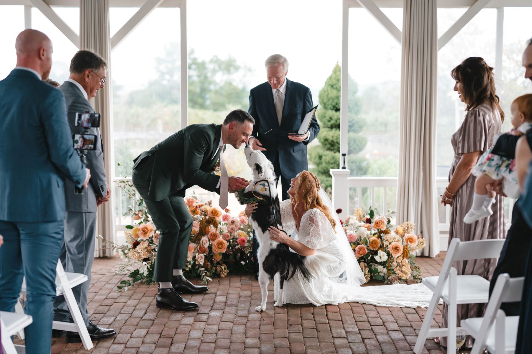 A couple at their wedding with their dog the woman is wearing a wedding dress and kneeling down to greet the dog who is greeting her partner who is wearing a black tuxedo as the guests watch and the officiant is in the background