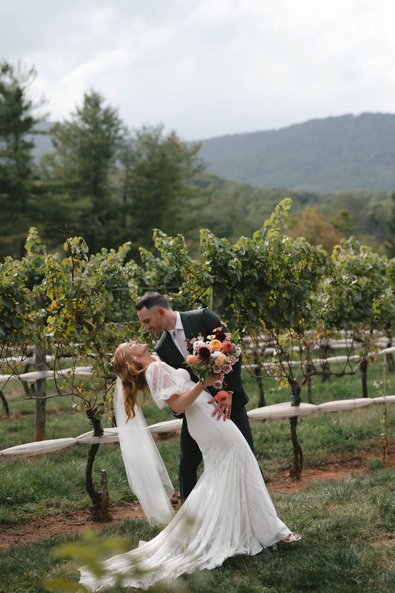 A couple in a vineyard the woman is wearing a white wedding dress and has red hair and is holding a bouquet of flowers and has a white veil on and her partner who is weraing a black tuxedo is dipping her backwards 