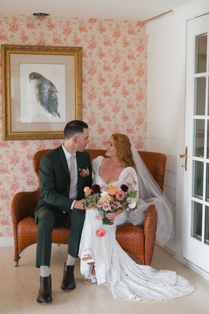 A woman in a white wedding dress, with a white veil and white heels is sitting on a wicker couch with her partner who is sitting next to hear and looking at her and is wearing a black tuxedo 